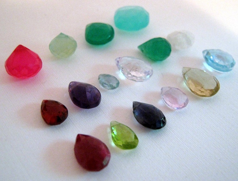 Puede incluir: Una colecci&oacute;n de gemas facetadas en varias formas y colores, incluyendo rojo, verde, morado y azul. Las gemas est&aacute;n dispersas sobre una superficie blanca, mostrando sus diferentes cortes y claridad.