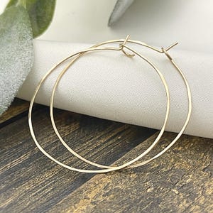 May include: A pair of gold-colored hoop earrings. The earrings are thin and circular, with a simple clasp closure. The hoops are displayed on a white cloth against a dark wooden surface.