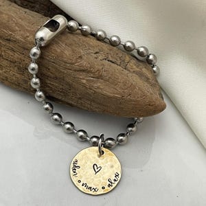 May include: A silver chain bracelet with a gold charm. The charm is round and has the names "alex", "max", and "niden" engraved on it, along with a small heart.