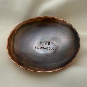 May include: A round, hammered copper dish with a dark, burnished finish. The center is stamped with the letters T and R separated by a heart symbol, and the date 8-22-2026 with heart symbols between the numbers.
