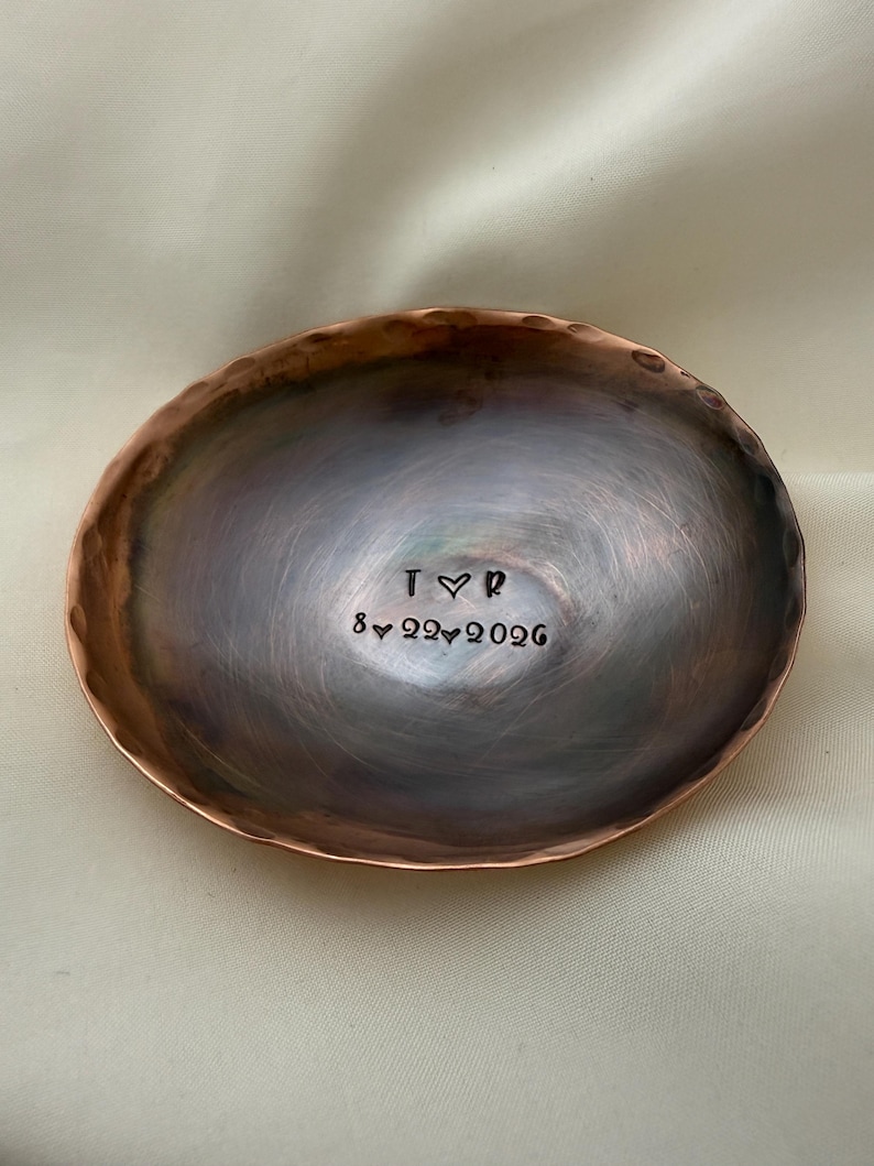 May include: A round, hammered copper dish with a dark, burnished finish. The center is stamped with the letters T and R separated by a heart symbol, and the date 8-22-2026 with heart symbols between the numbers.