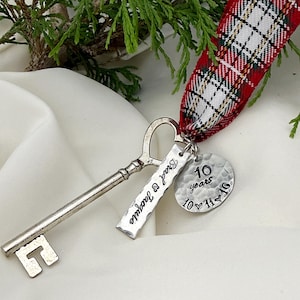 Puede incluir: Un adorno de llave antigua de metal plateado unido a una cinta de cuadros roja, blanca y verde. Dos dijes de metal cuelgan de la llave: una etiqueta rectangular grabada con Brad & Jacquie y un disco martillado con 10 years 10-11-16.