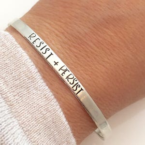 May include: Silver bracelet with the words "Resist + Persist" engraved on it.