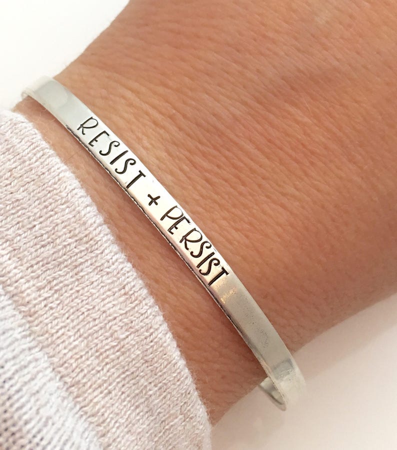 May include: Silver bracelet with the words "Resist + Persist" engraved on it.