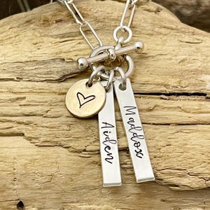 May include: Silver chain necklace with a toggle clasp and two silver bar charms engraved with the names "Aiden" and "Maddox". A small gold heart charm is also attached to the necklace.