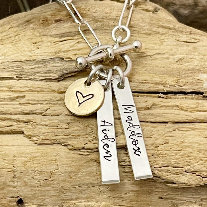May include: Silver chain necklace with a toggle clasp and two silver bar charms engraved with the names "Aiden" and "Maddox". A small gold heart charm is also attached to the necklace.