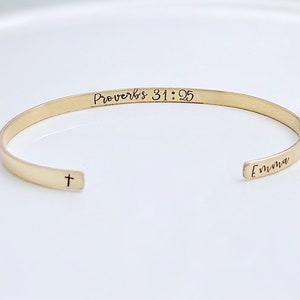 May include: A gold-colored cuff bracelet with the text "Proverbs 31:25" and the name "Emma" engraved on it. A small cross is engraved on the opposite end of the bracelet. The bracelet is displayed on a white surface.