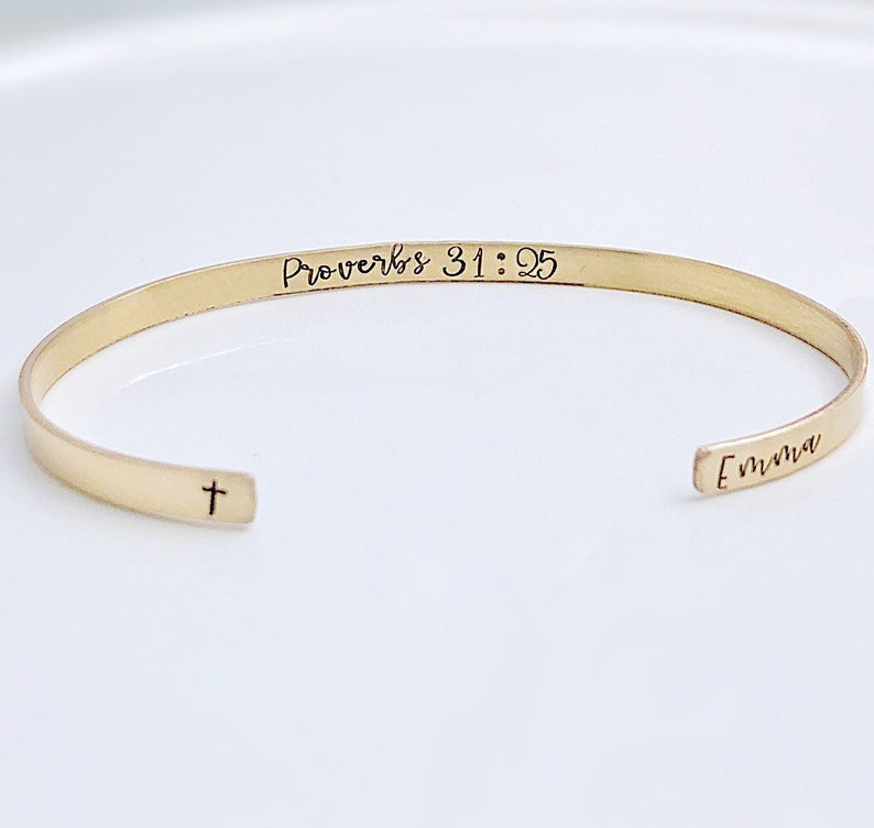 May include: A gold-colored cuff bracelet with the text "Proverbs 31:25" and the name "Emma" engraved on it. A small cross is engraved on the opposite end of the bracelet. The bracelet is displayed on a white surface.