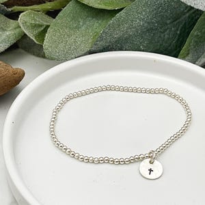 May include: A silver beaded bracelet with a small, round charm featuring a black cross. The bracelet is displayed on a white, shallow dish, with green foliage in the background.