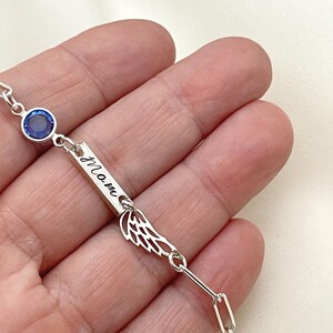 Op de afbeelding: Zilveren armband met een blauwe kristal, een staafvormige bedel met de gravure "Mom" en een vleugelbedel.