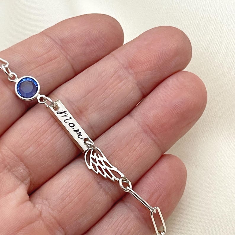 Op de afbeelding: Zilveren armband met een blauwe kristal, een staafvormige bedel met de gravure "Mom" en een vleugelbedel.