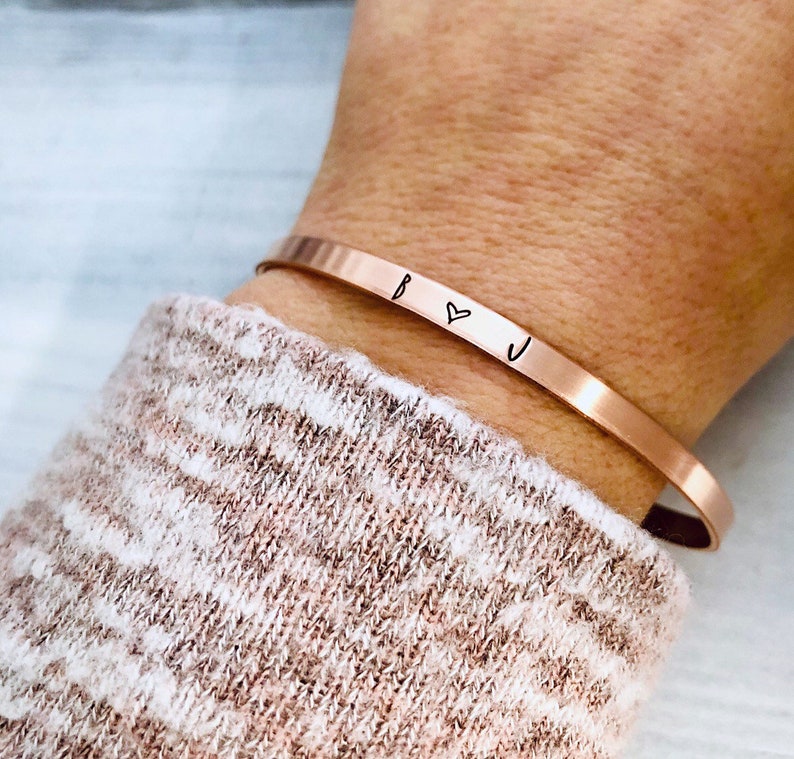 May include: A rose gold cuff bracelet with the initials "B" and "J" separated by a heart symbol.