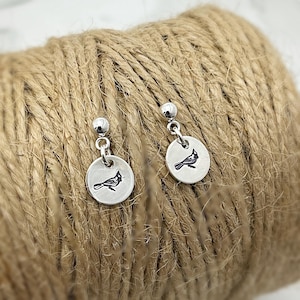 May include: A pair of silver-colored earrings. Each earring features a small, round charm with a black cardinal bird design. The earrings are dangling from a small silver ball stud. The earrings are displayed on a brown, textured surface.