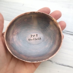 May include: A round, hammered copper bowl with a dark, weathered patina. The interior is stamped with the text J heart B and the date 10 heart 11 heart 03 in black ink. The bowl has a rustic, uneven edge and a polished copper rim.