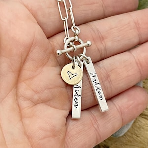 May include: Silver chain necklace with a toggle clasp and three charms. The charms are a small round charm with a heart engraved on it, and two rectangular charms with the names "Aiden" and "Maddox" engraved on them.