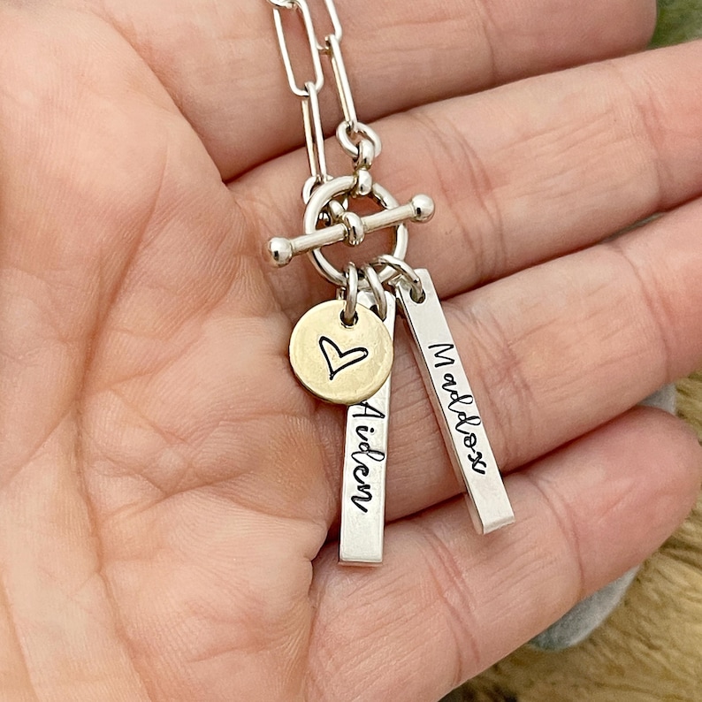 May include: Silver chain necklace with a toggle clasp and three charms. The charms are a small round charm with a heart engraved on it, and two rectangular charms with the names "Aiden" and "Maddox" engraved on them.
