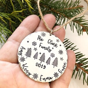 May include: Silver Christmas ornament with the text "The Clark Family 2019" and the names "Amy", "Kenny", "Emma", and "Gavin" around the edge. The ornament features a design of four stylized pine trees and snowflakes.