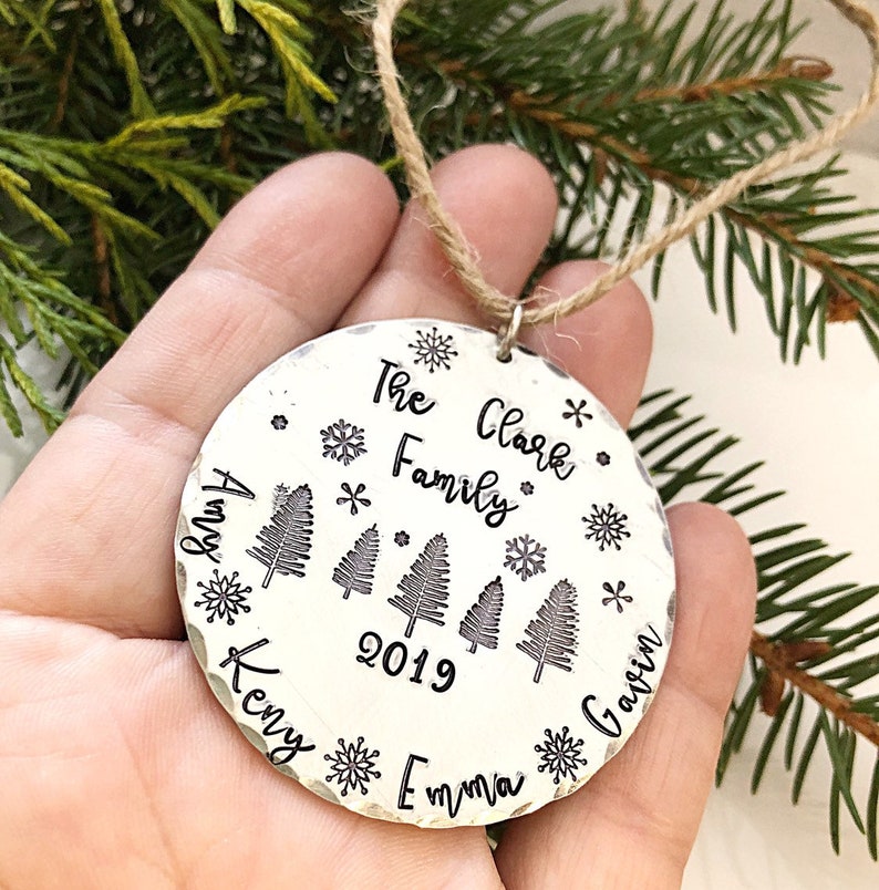 May include: Silver Christmas ornament with the text "The Clark Family 2019" and the names "Amy", "Kenny", "Emma", and "Gavin" around the edge. The ornament features a design of four stylized pine trees and snowflakes.