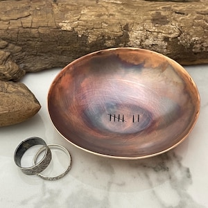 May include: A handmade copper bowl with a textured finish and a tally mark design. Two silver rings sit beside the bowl. The bowl's interior has a gradient of colours, from copper to purple.