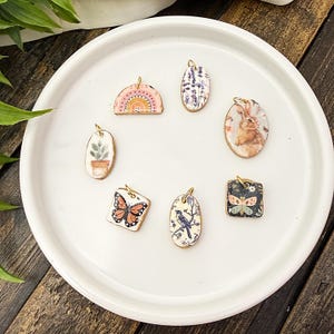 May include: A collection of seven small, decorative pendants with gold-colored loops. The pendants feature various designs, including a rainbow, a bunny, a butterfly, a bird, and a potted plant. The pendants are arranged on a white plate.