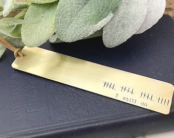 May include: A brass bookmark with a leather cord. The bookmark features the text "I still do" and tally marks. It rests on a dark blue book, with green foliage in the background. The bookmark is rectangular.
