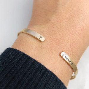 May include: Gold-toned cuff bracelet with a cross and the name "Emma" engraved on the ends. The open design allows for easy wear. The bracelet is shown on a wrist with a dark blue sweater.