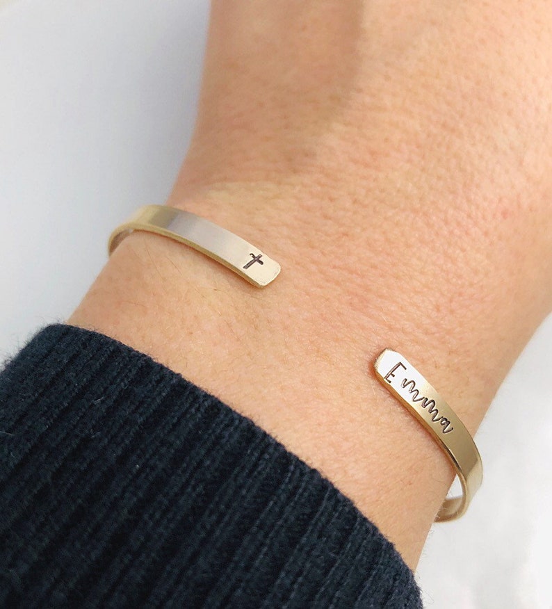 May include: Gold-toned cuff bracelet with a cross and the name "Emma" engraved on the ends. The open design allows for easy wear. The bracelet is shown on a wrist with a dark blue sweater.