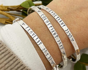 May include: Three silver bracelets with hammered textures. Each bracelet has a heart symbol and a bible verse engraved on it. The verses are Proverbs 3:21, Colossians 3:12, and Ephesians 3:20.