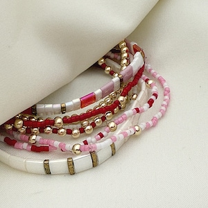 May include: A collection of beaded bracelets in shades of red, pink, white, and gold. The bracelets feature small beads and larger gold-colored beads. Some bracelets have rectangular white beads with gold accents. The bracelets are displayed on a white surface.