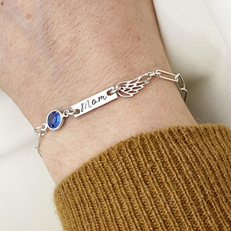 Op de afbeelding: Zilveren armband met een blauwe kristallen bedel, een klein gegraveerd label met de tekst "mom" en een delicate vleugelbedel.