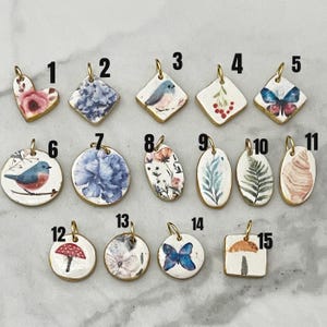 May include: A collection of fifteen ceramic pendants with gold-colored edges. The pendants feature various designs, including flowers, birds, butterflies, and mushrooms. Shapes vary from hearts and diamonds to circles and squares. Each pendant has a small loop for hanging.z
