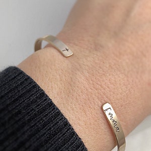 May include: A gold-colored cuff bracelet with a small cross engraved on one end and the name "Emma" engraved on the other. The bracelet is worn on a wrist, with a black sweater in the background.