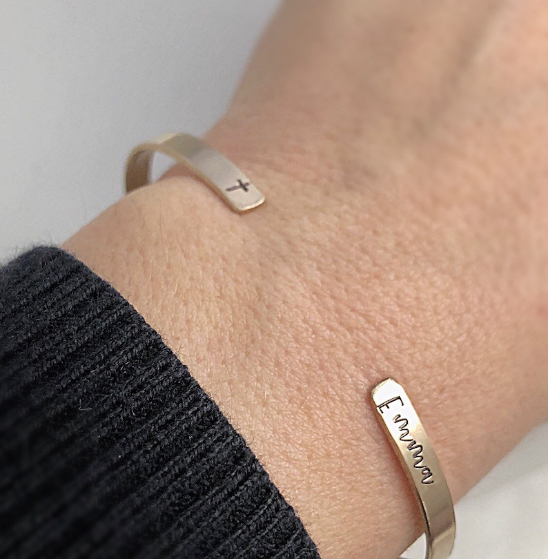 May include: A gold-colored cuff bracelet with a small cross engraved on one end and the name "Emma" engraved on the other. The bracelet is worn on a wrist, with a black sweater in the background.