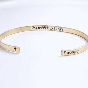May include: A gold-colored cuff bracelet with the text "Proverbs 31:25" and the name "Emma" engraved on it. A small cross is engraved near the opening of the bracelet. The bracelet is on a white surface.