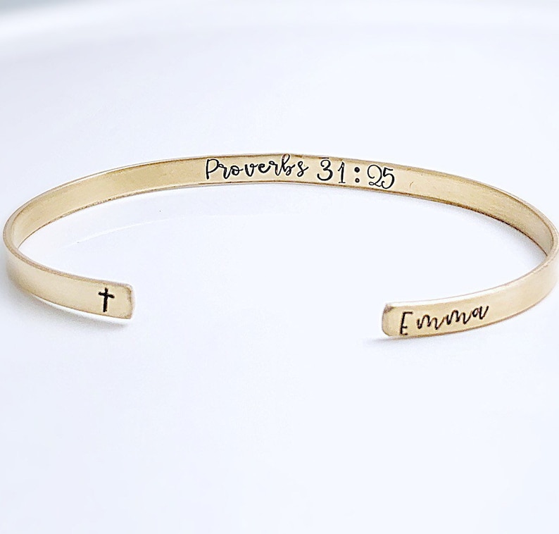May include: A gold-colored cuff bracelet with the text "Proverbs 31:25" and the name "Emma" engraved on it. A small cross is engraved near the opening of the bracelet. The bracelet is on a white surface.