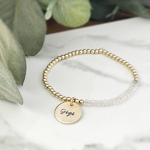 May include: Gold beaded bracelet with a round charm engraved with the name "Gigi". The bracelet features a section of clear, faceted beads. The charm and beads are gold in color.