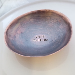 May include: A small, oval-shaped copper trinket dish with a hammered texture and a darkened, oxidized finish. The center is hand-stamped with the letters J, a heart symbol, and B, followed by the date 10, a heart, 11, a heart, and 03.