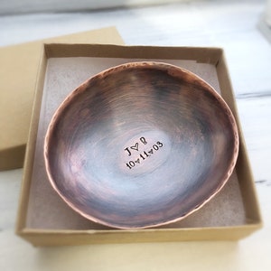 May include: A round, hammered copper bowl with a dark, weathered patina finish. The interior is stamped with the text J heart B and the date 10 heart 11 heart 03. The bowl is presented inside a square, light brown cardboard gift box.