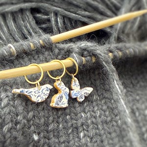 May include: Close-up of knitting needles with three decorative stitch markers. The markers are gold-toned rings with ceramic charms shaped like a bird, a bunny, and a butterfly. Each charm has a blue and white floral pattern. The yarn is gray.