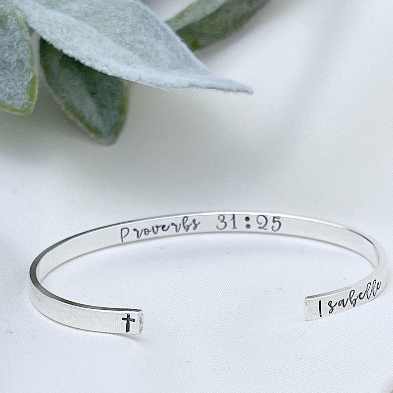 May include: A silver cuff bracelet with the inscription "Proverbs 31:25" and the name "Isabelle". The bracelet features a small cross symbol and is displayed on a white surface.