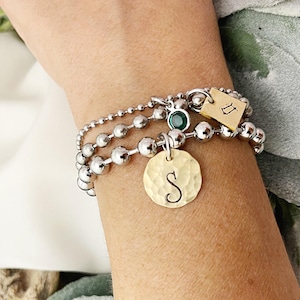 May include: A set of three silver beaded bracelets with personalized charms. One charm is a hammered gold disc with the letter "S", another is a small gold tag with a zodiac symbol, and a third has a green gemstone.