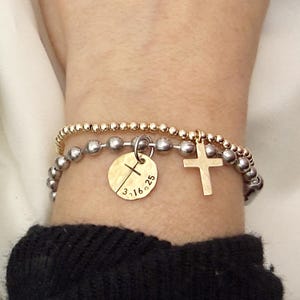 May include: Two beaded bracelets, one gold and one silver, are stacked on a wrist. The silver bracelet has a round charm with a cross and the date 3.16.25. A gold cross charm hangs from the gold bracelet.