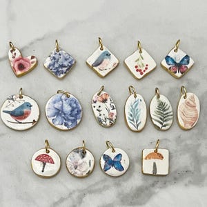 May include: A collection of handmade ceramic pendants with gold-colored edges. The pendants feature various designs, including flowers, birds, butterflies, and mushrooms. Each pendant has a small gold loop for hanging.