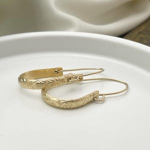 May include: A pair of gold-colored hoop earrings with an engraved floral design. The earrings are on a white surface. The earrings have a hook closure.