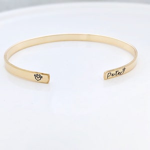 May include: Gold-toned bangle bracelet with a paw print and heart design on one side and the name "Petey" engraved on the other side.