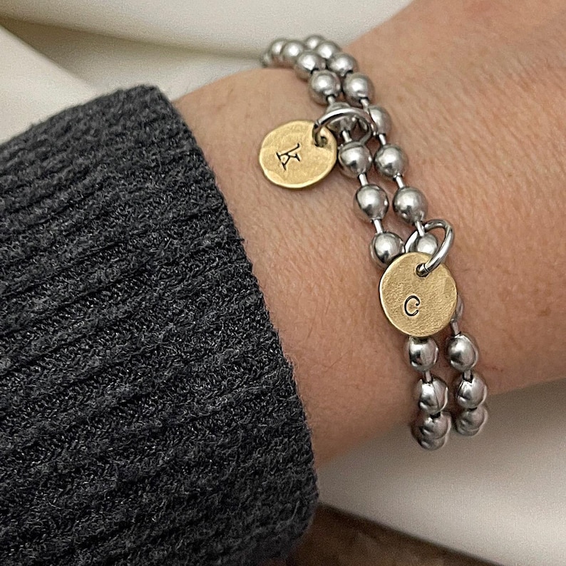 May include: A silver beaded bracelet with two gold-colored circular charms. One charm has the letter "K" and the other has the letter "C". The bracelet is worn on a wrist next to a gray sweater.