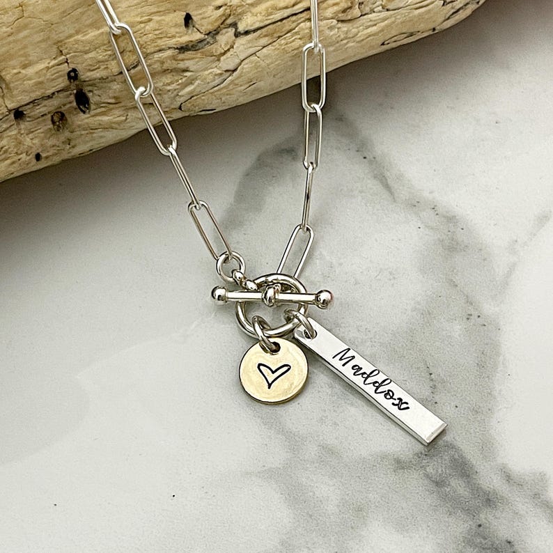 May include: A silver chain necklace with a toggle clasp. The necklace has two charms: a round charm with a heart engraved on it and a rectangular charm with the name "Maddox" engraved on it.