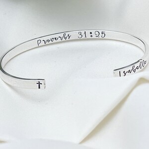 May include: A silver-colored cuff bracelet with the inscription "Proverbs 31:25" and the name "Isabelle". A small cross is etched on the bracelet's edge. The bracelet is displayed on a white, textured surface.