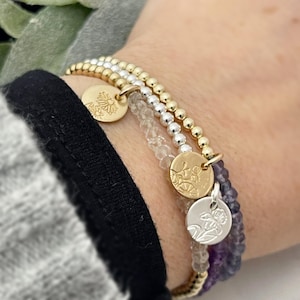 May include: Three gold and silver beaded bracelets with floral charms. The bracelets are stacked on a person's wrist.