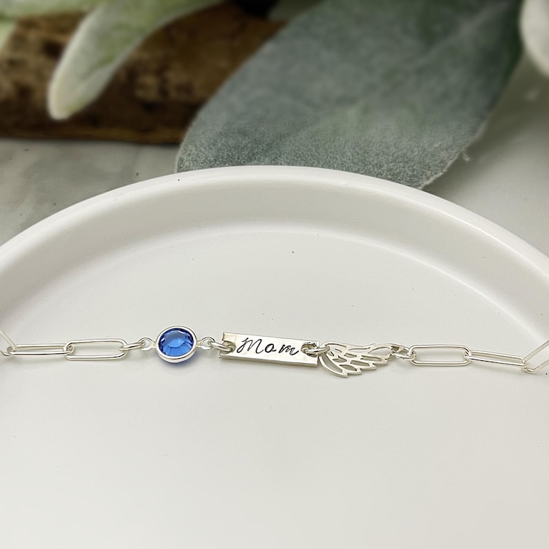 Op de afbeelding: Een zilveren kettingarmband met een blauwe kristallen bedel, een kleine zilveren vleugelbedel en een zilveren staafbedel met de gravure "Mom".